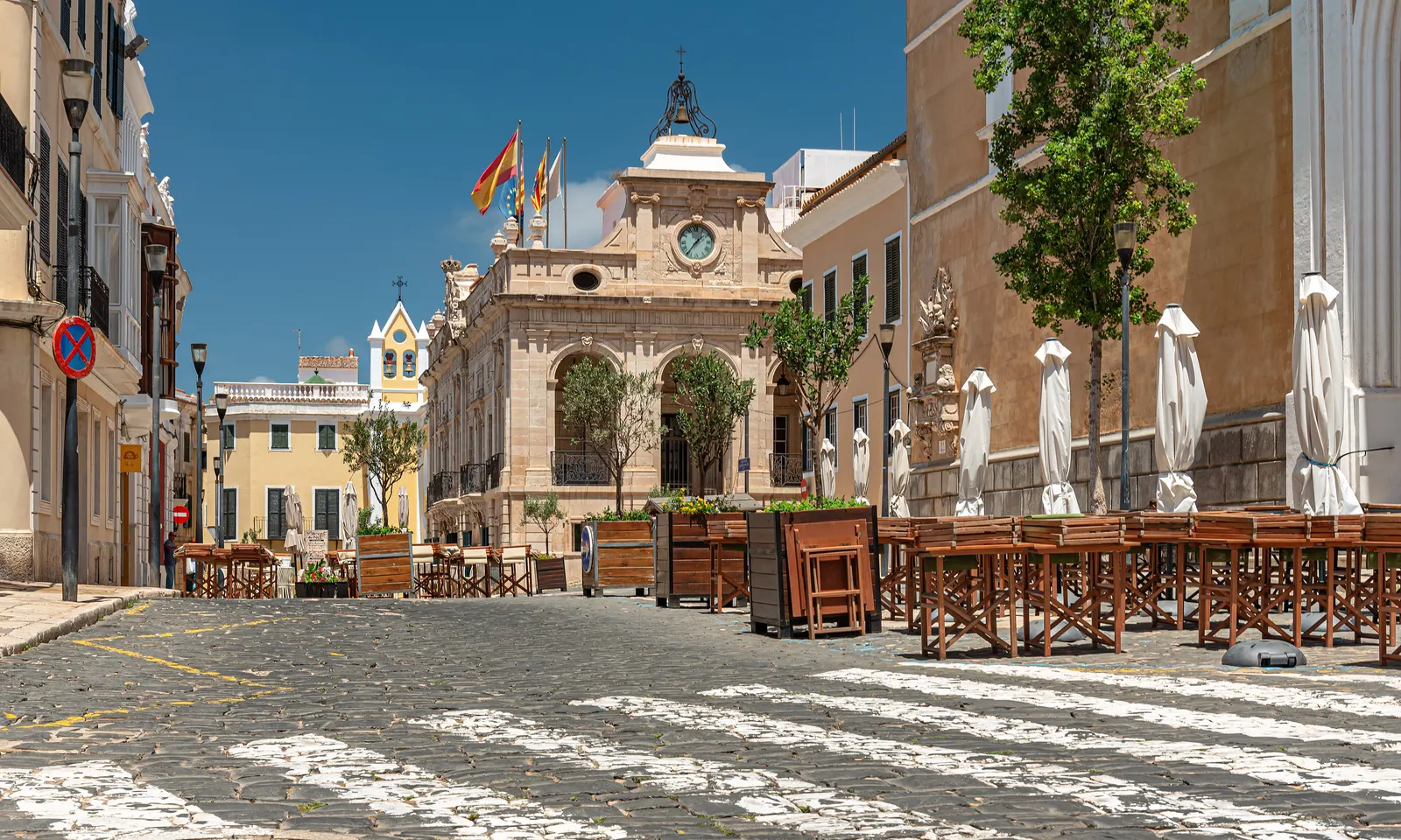 La plaza del Ayuntamiento de Maó, con su fachada de piedra clara, sus terrazas recogidas y una torre con reloj al fondo.