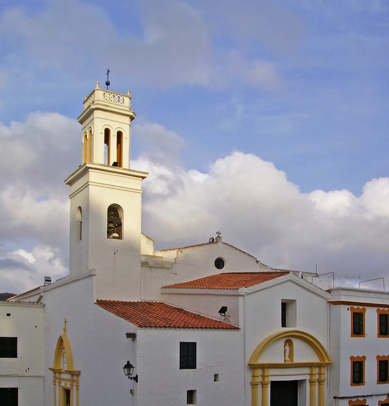 El campanario y la fachada de la iglesia de Sant Bartomeu, en el centro del pueblo de Ferreries, contra un cielo con nubes.