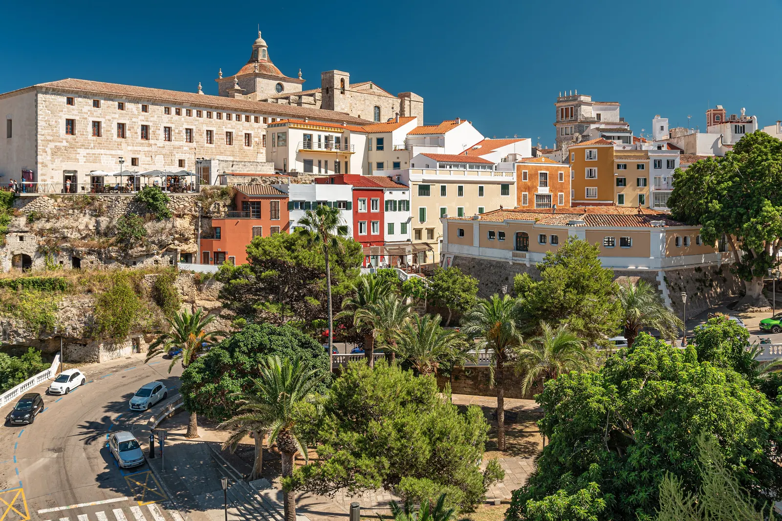 El perfil del casco antiguo de Maó sobre el puerto, con la Iglesia del Carme, casas de colores y palmeras asomando entre los tejados.