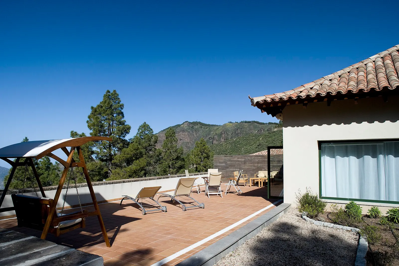 Terraza soleada con tumbonas y un balancín junto a una casa de tejado de teja, con pinos y montañas de la cumbre al fondo.