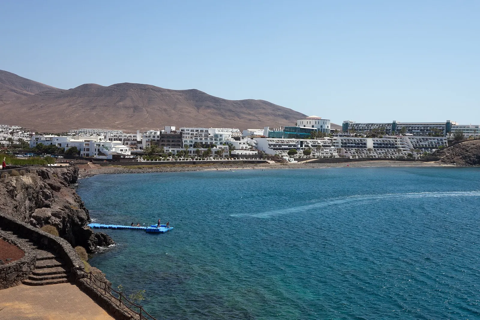 Una cala de agua turquesa en Playa Blanca, con hoteles bajos y blancos al pie de la montaña.