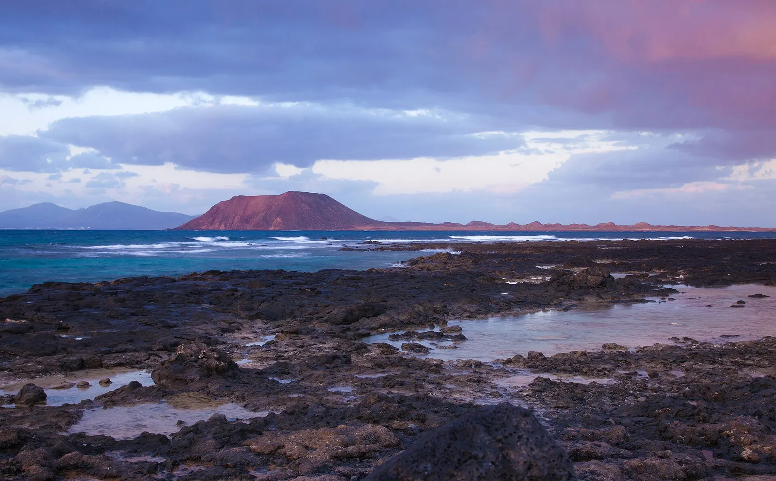 El islote de Lobos al atardecer, con su volcán recortado sobre el mar y una costa de roca negra con charcos en primer plano.