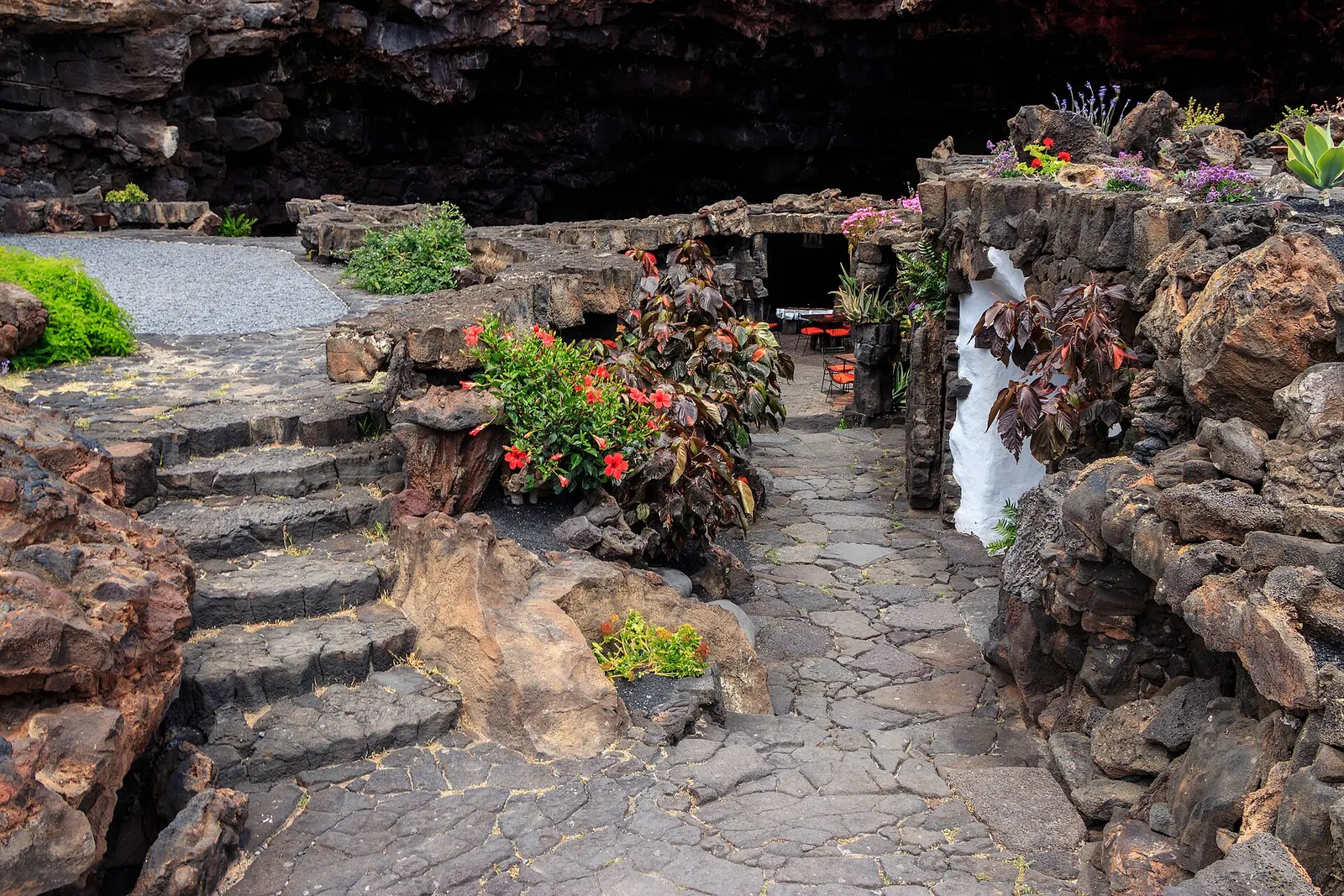 Una escalera de piedra volcánica baja entre plantas y flores hacia la boca oscura del tubo de lava.