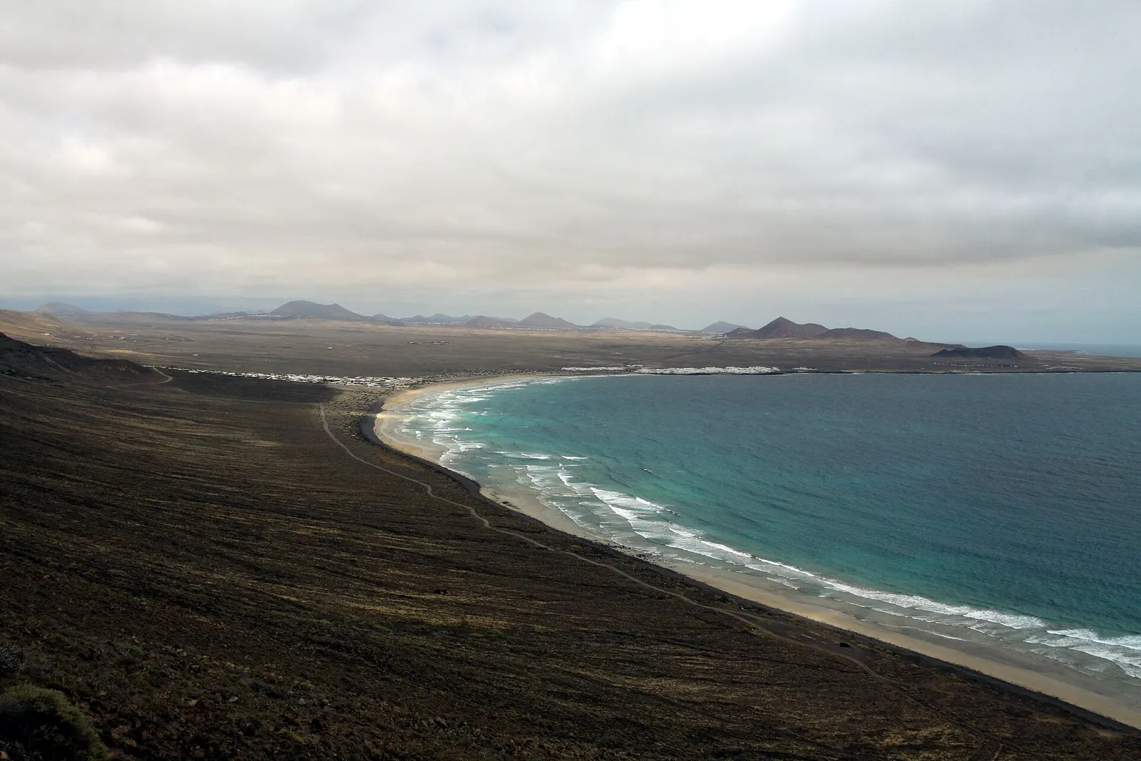 La larga playa de Famara curva bajo un cielo nublado, con el Atlántico verde azulado rompiendo en la orilla.