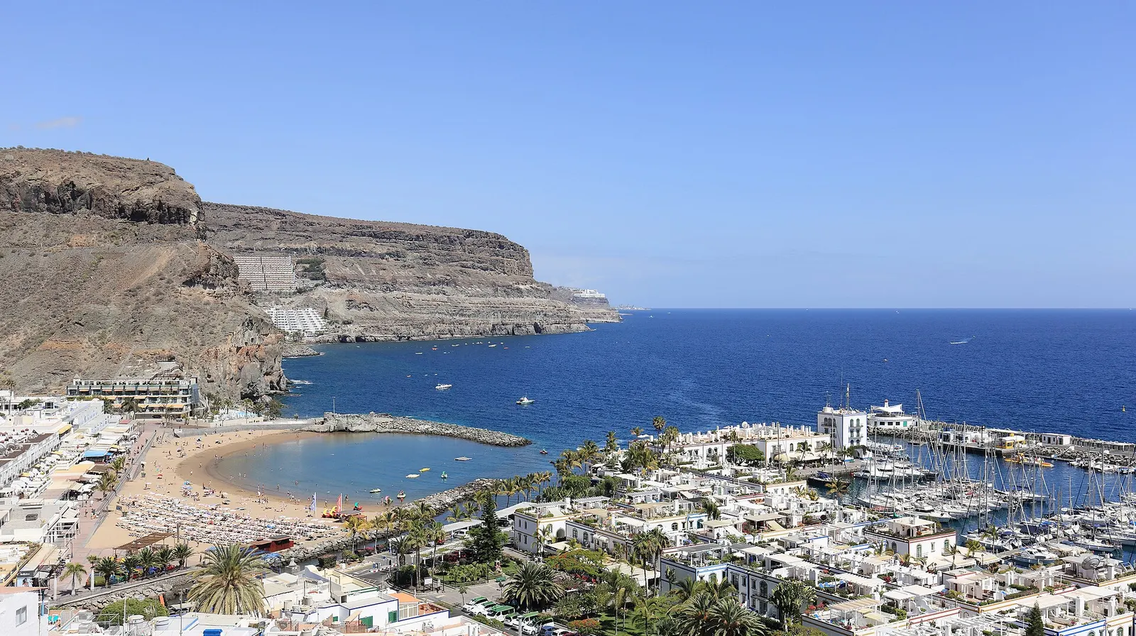 Puerto de Mogán desde lo alto: el puerto deportivo lleno de veleros, la playa y los acantilados sobre un mar azul intenso.