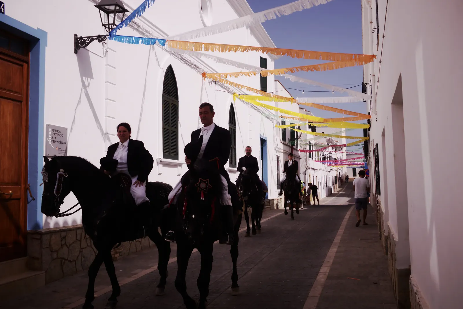 Dos caballeros de fiesta a caballo, de negro y con sombrero, bajan por una calle blanca de Es Mercadal bajo guirnaldas de banderines de colores