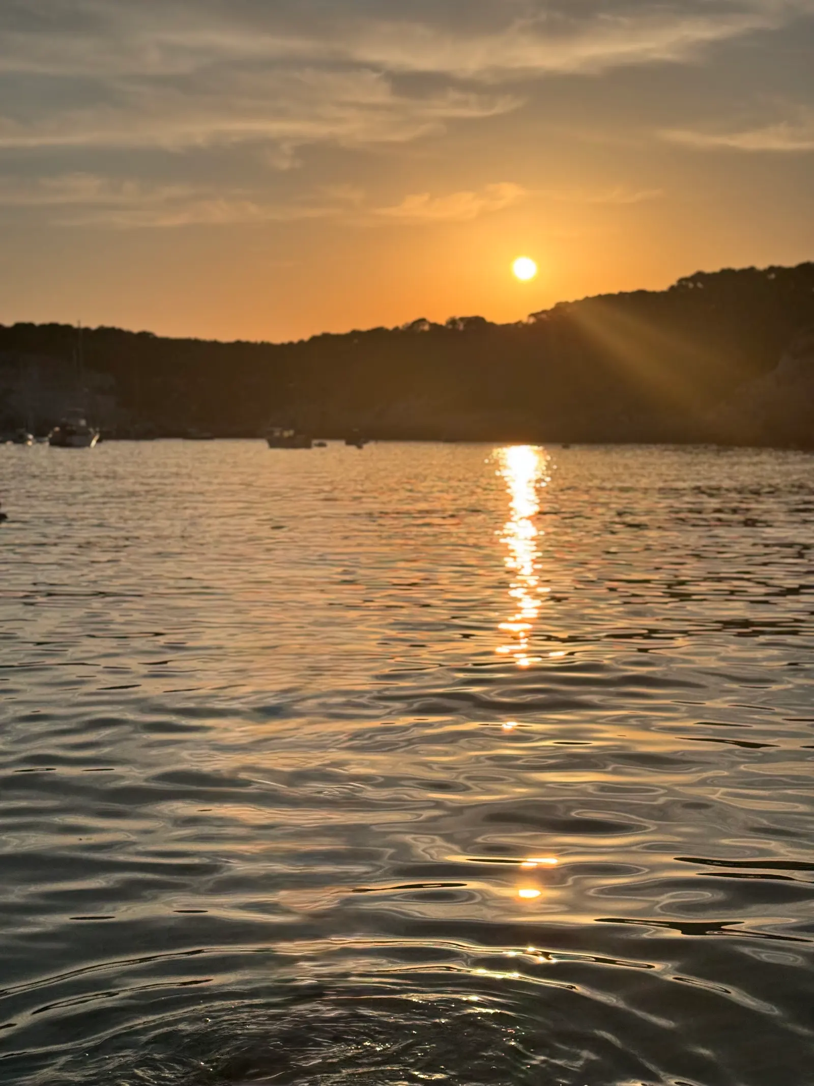 Un atardecer sereno en una cala de Menorca: el sol muy bajo sobre el agua en calma, con dos barcas fondeadas en silueta.