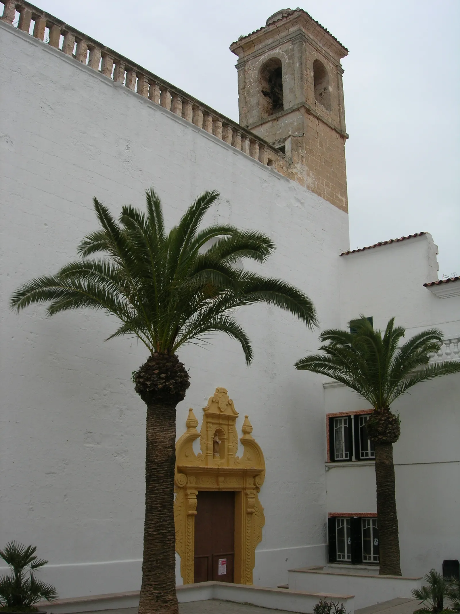 La facciata bianca del convento di Sant Dídac, nel centro di Alaior, con il portale barocco ocra, il campanile e due palme davanti.