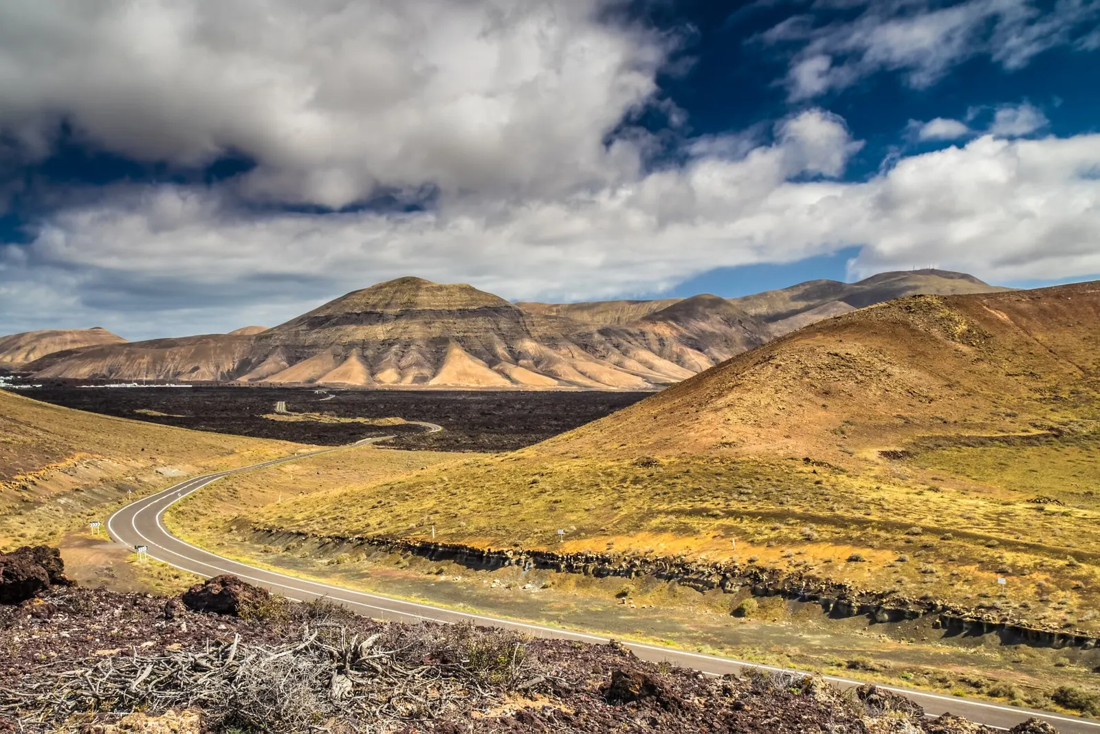 Una carretera serpentea entre campos de lava y montañas ocres bajo un cielo de nubes blancas.