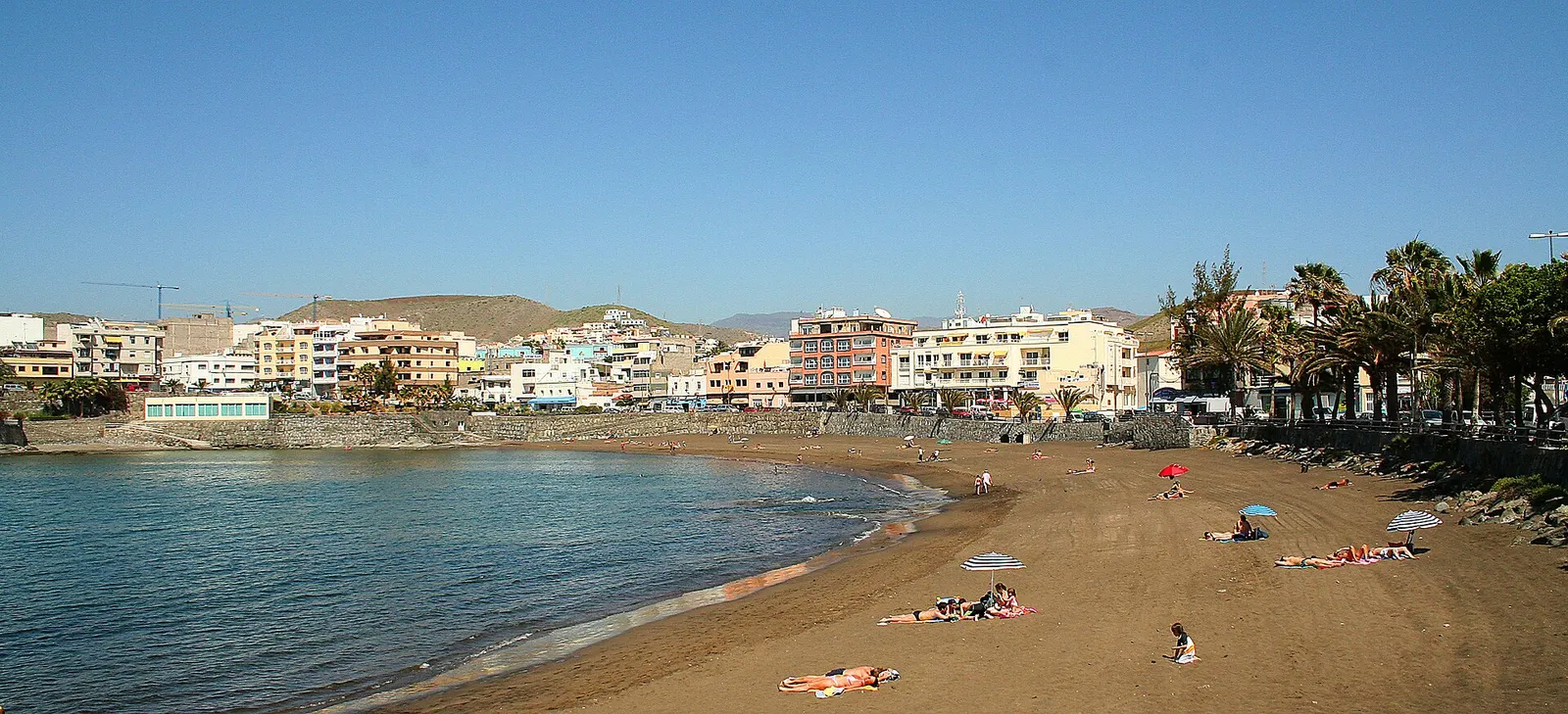 La playa de Arguineguín con agua tranquila, sombrillas dispersas y el pueblo de casas bajas tras las palmeras.