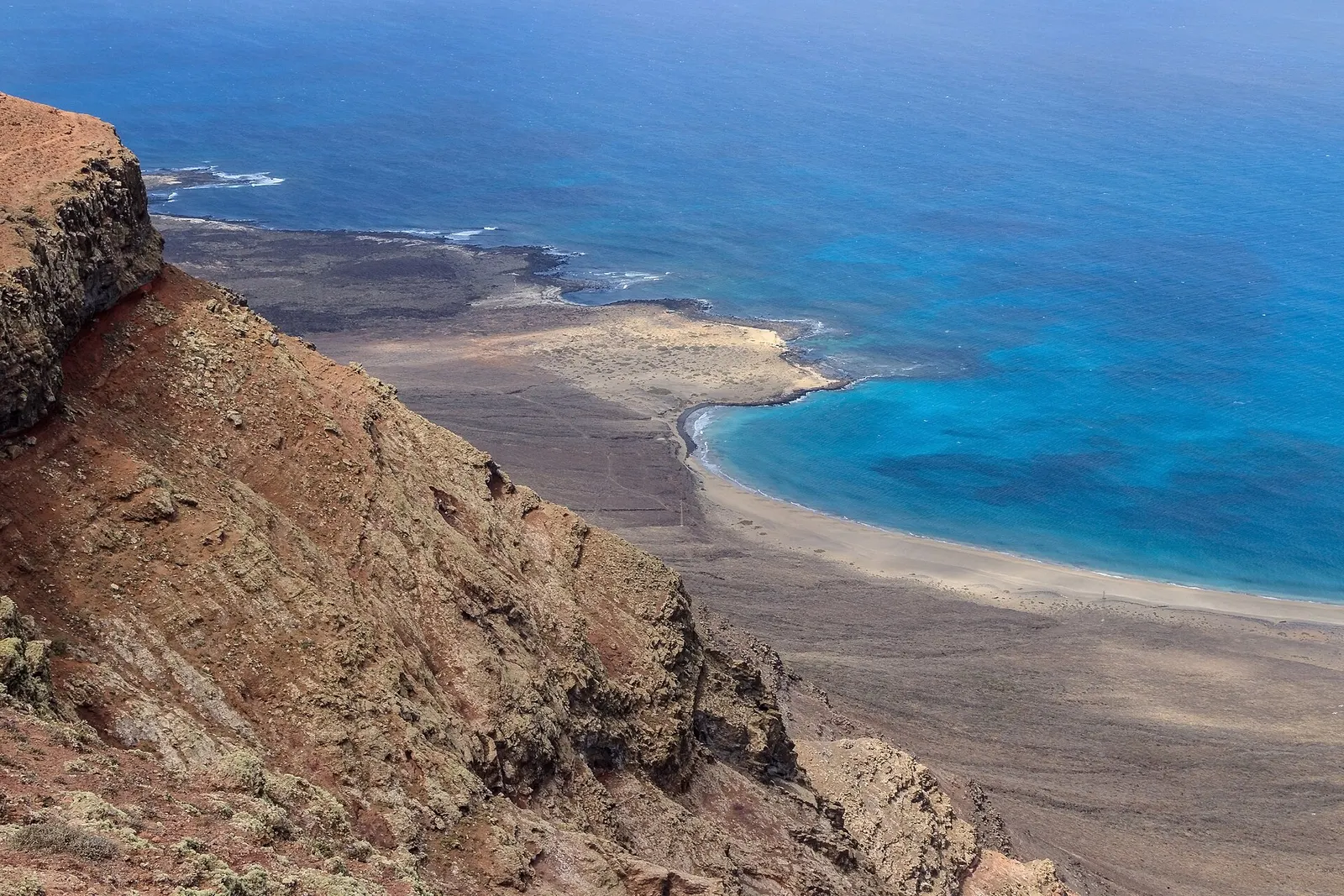 Desde lo alto del risco junto al Mirador del Río, la ladera cae hacia una playa de arena clara y un mar turquesa.