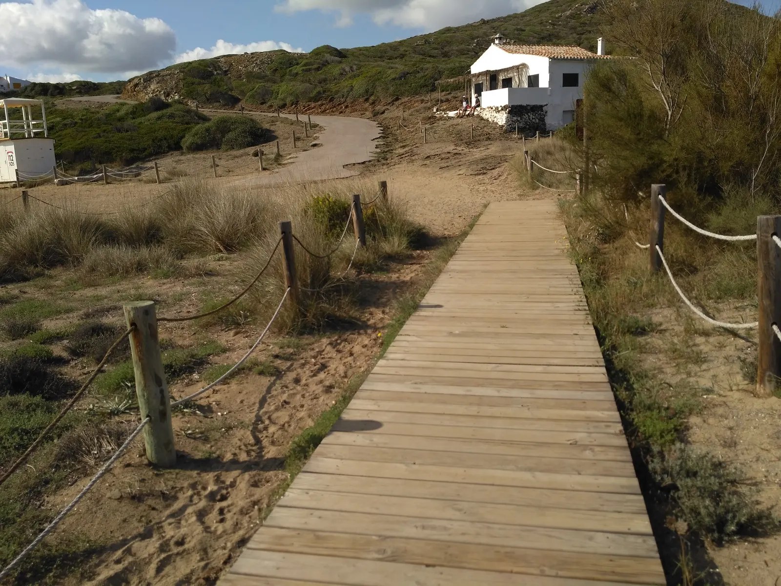 Una passerella in legno del Camí de Cavalls lungo la costa, vicino a Sa Mesquida, con una casa bianca sullo sfondo tra la macchia mediterranea.