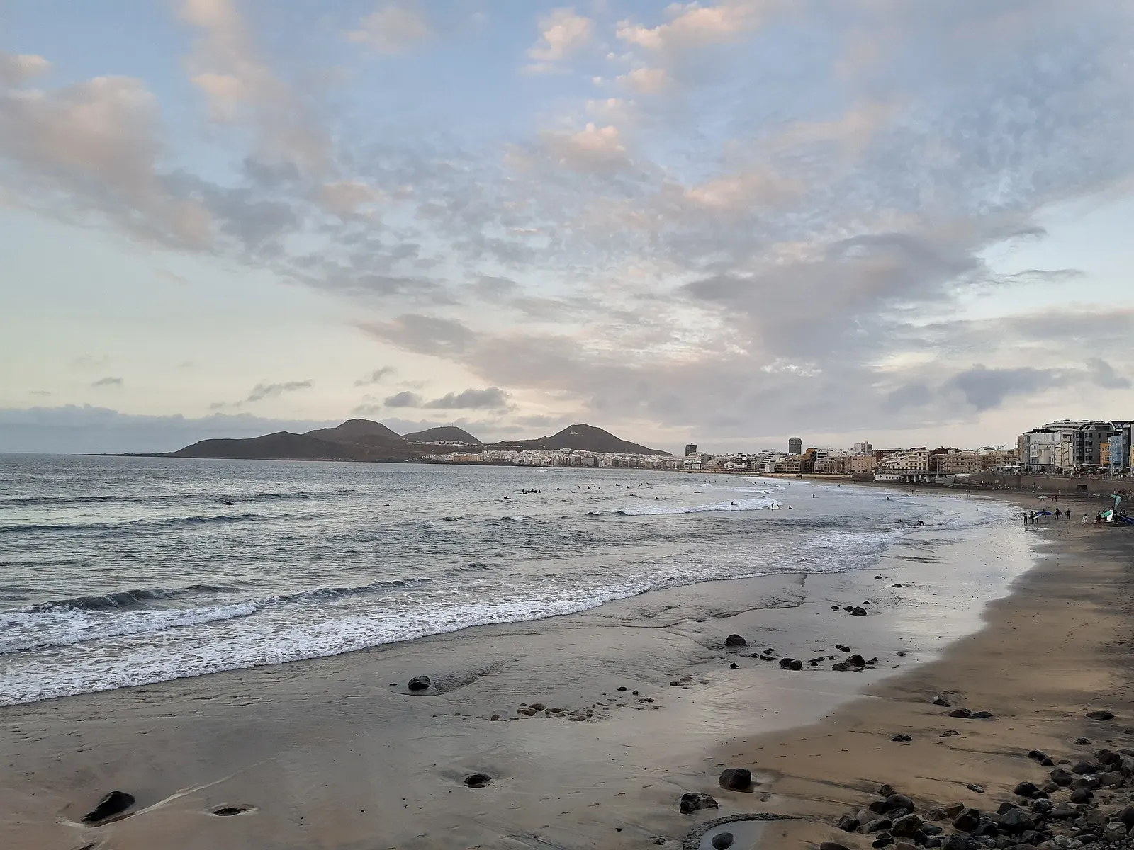 Las Canteras al atardecer desde el sur, con la orilla mojada, olas suaves y las montañas de La Isleta bajo un cielo rosado.