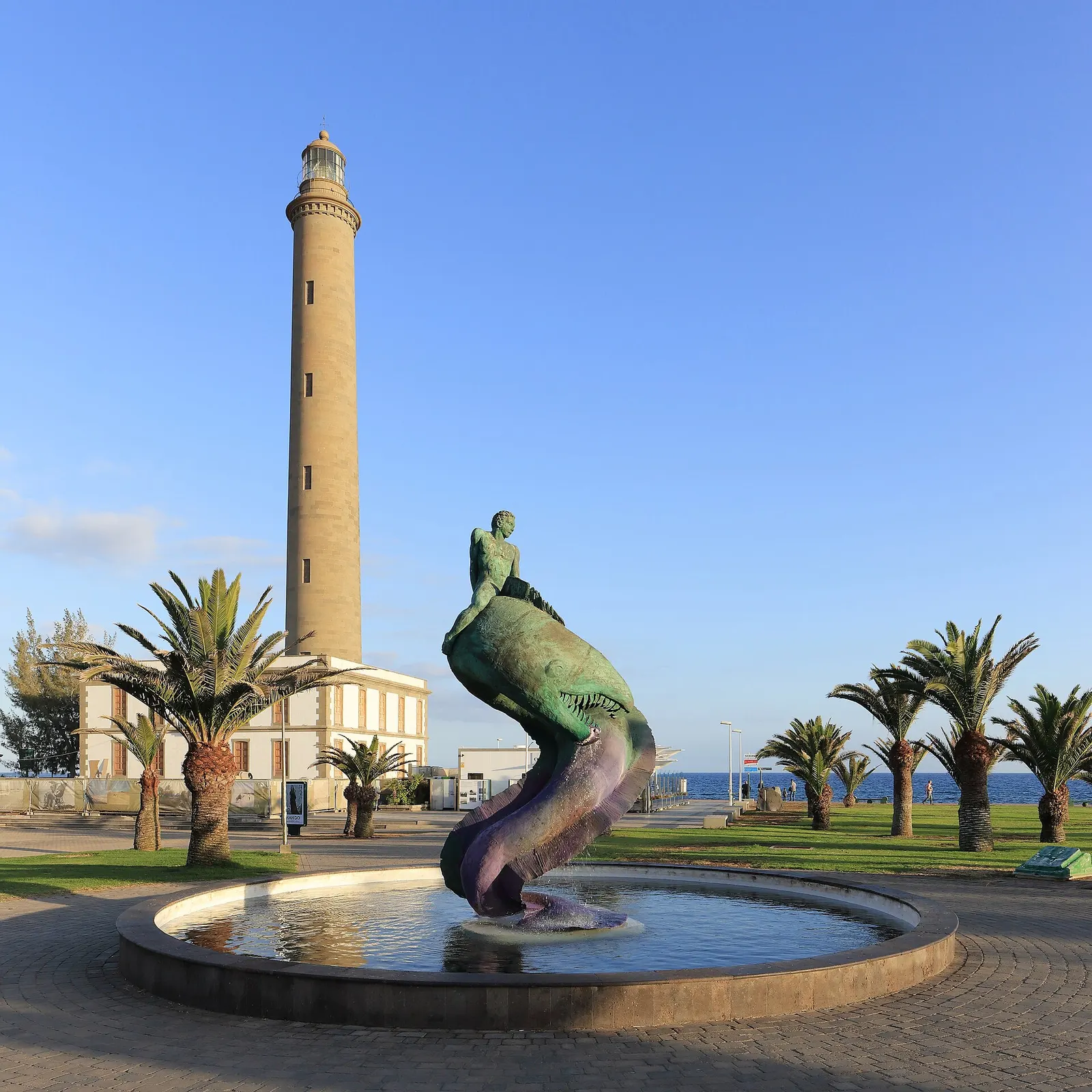 Der Leuchtturm von Maspalomas am frühen Tag, mit dem Meerjungfrauenbrunnen, Palmen und der leeren Promenade unter klarem Himmel.