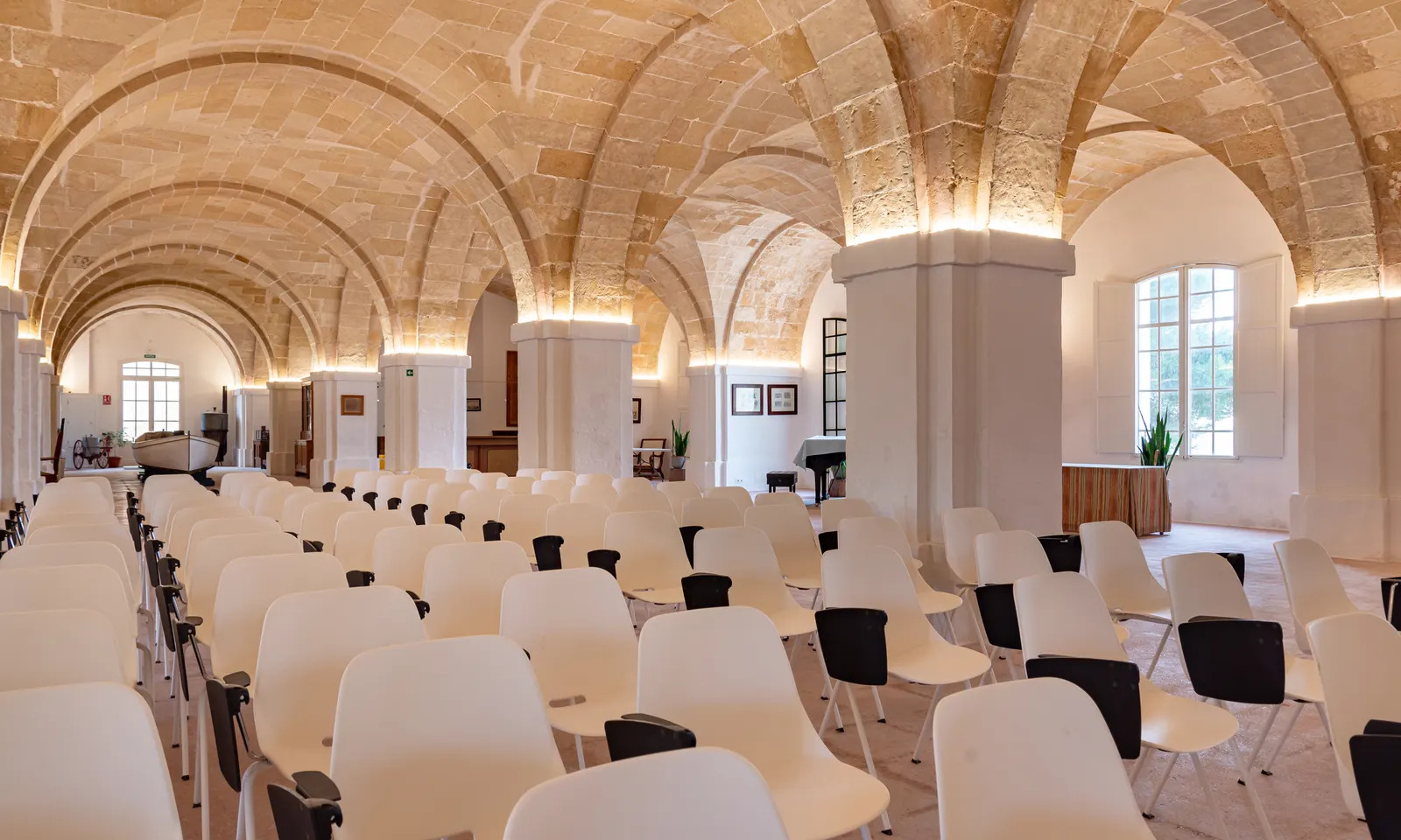 La sala voltata in pietra del Llatzeret de Maó, allestita con file di sedie per un evento, con archi a tutto sesto e luce naturale dalle finestre.