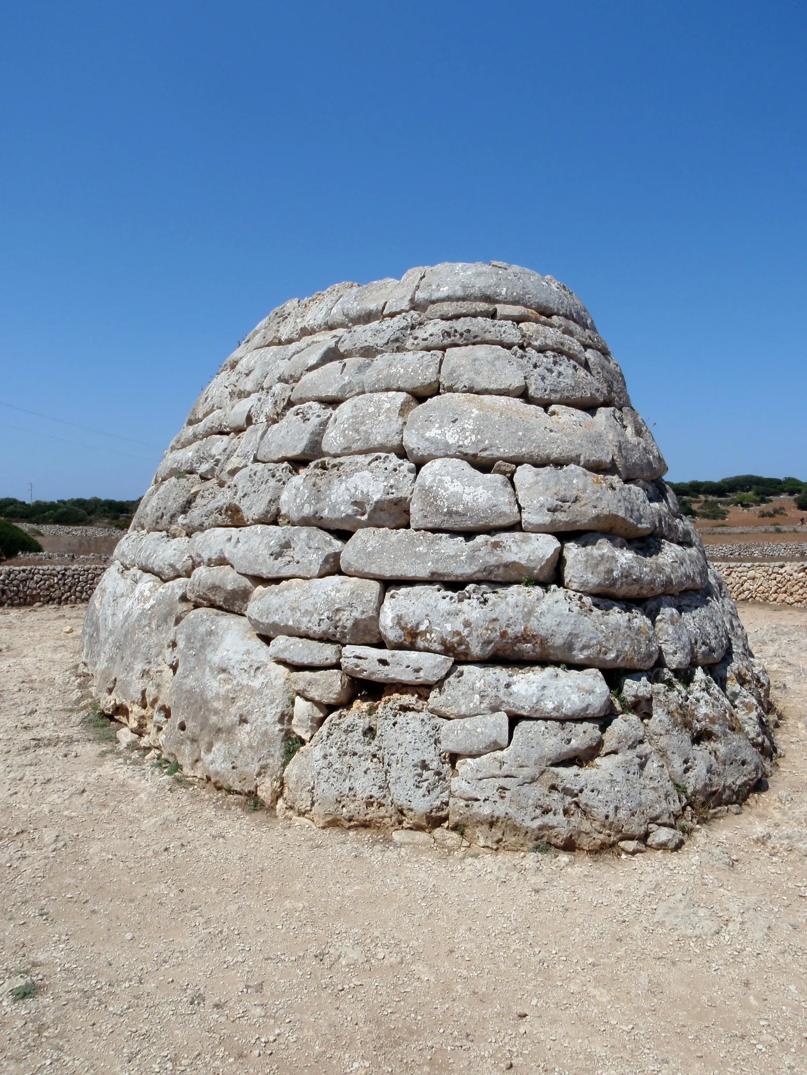 La Naveta des Tudons, sepolcro talaiotico a forma di nave rovesciata, sotto un cielo azzurro terso.