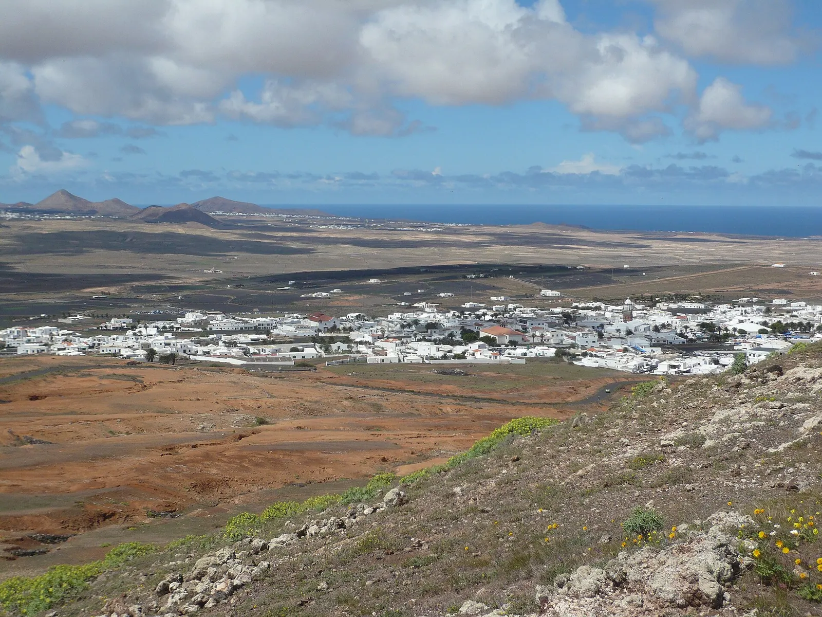 La villa blanca de Teguise en medio de la llanura, vista desde la ladera con el mar y los volcanes en el horizonte.