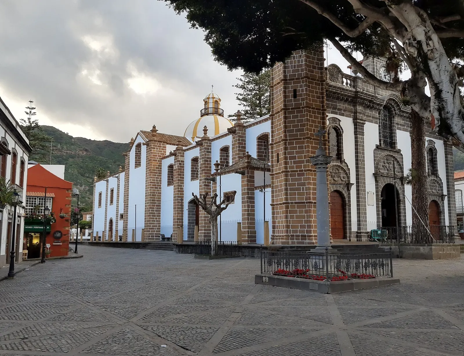 La basílica del Pino en Teror, blanca con torre de piedra, frente a la plaza empedrada y con los montes verdes detrás.