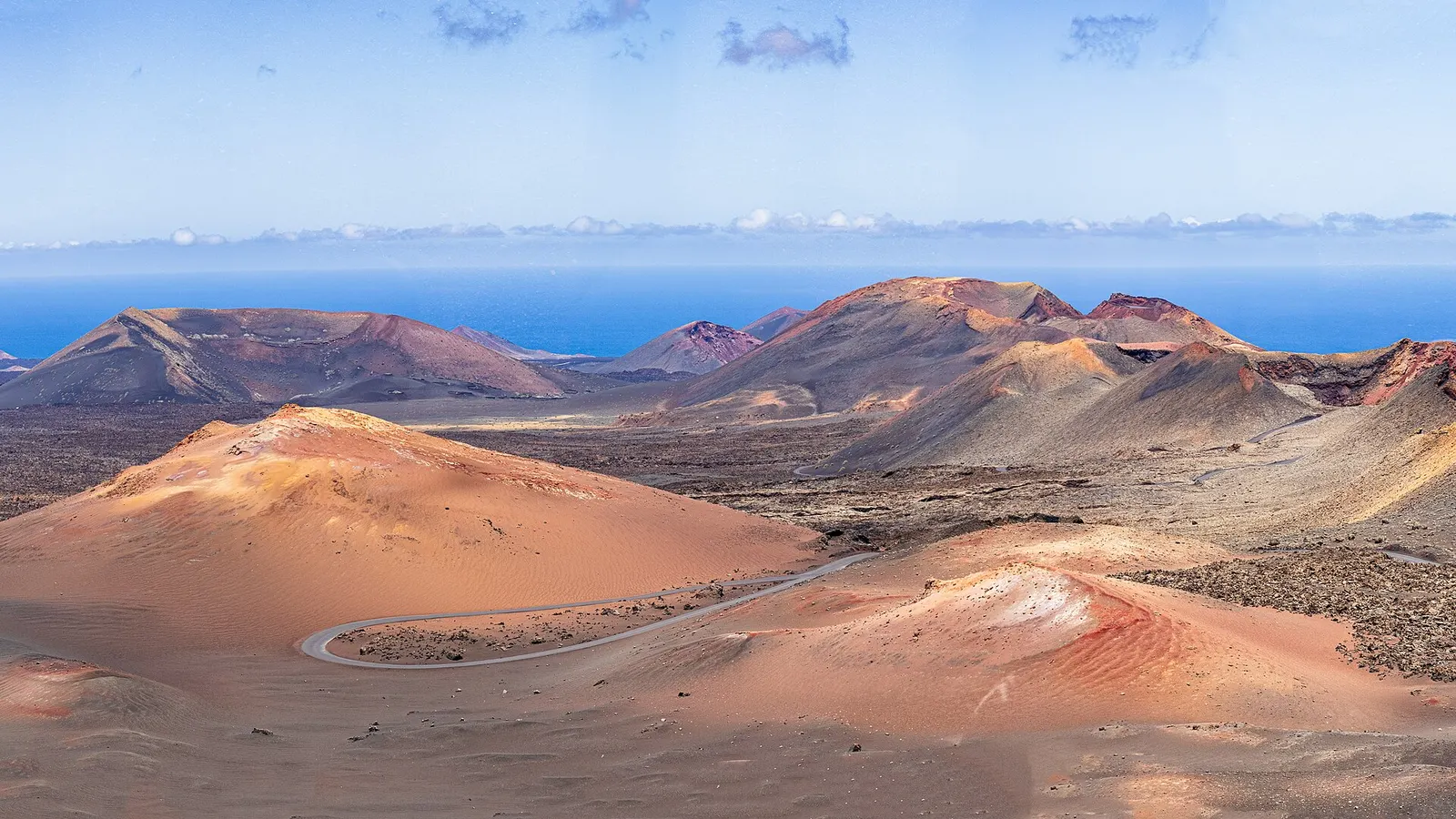 Conos volcánicos ocres y rojizos de Timanfaya, con la ruta asfaltada serpenteando entre ellos y el mar al fondo.