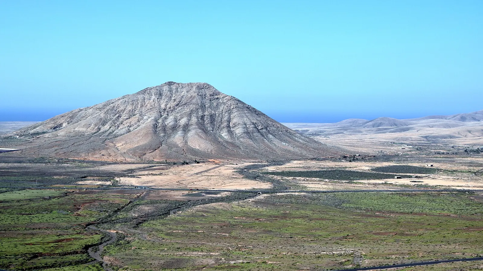 La montaña de Tindaya, solitaria y de laderas claras, se alza sobre la llanura verde del norte con el mar al fondo.