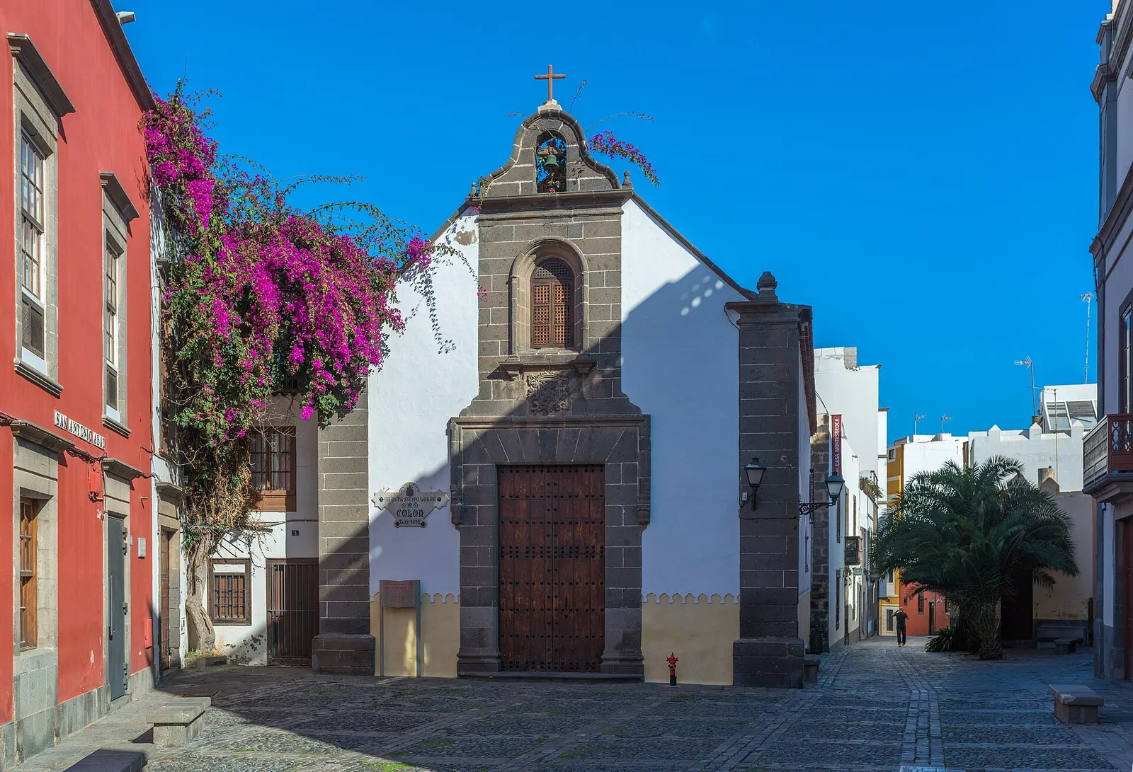 La ermita de San Antonio Abad en Vegueta, de piedra gris y cal, con una buganvilla fucsia y la calle empedrada vacía.