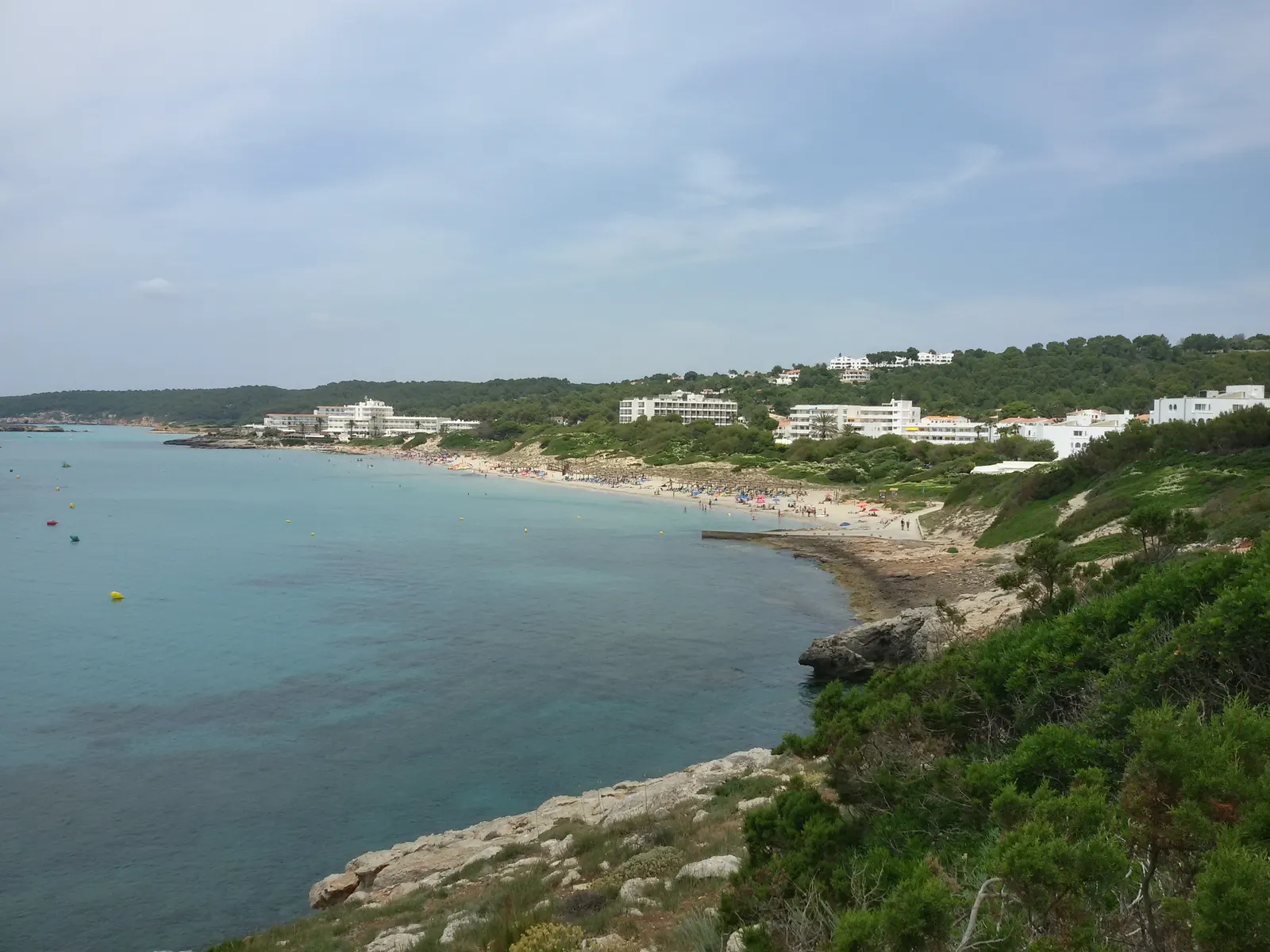 La bahía de Sant Tomàs desde un promontorio: agua turquesa, arena dorada y colinas de pinar al fondo.