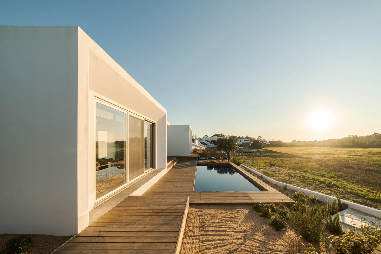 Casa blanca minimalista con piscina y terraza de madera frente a un paisaje mediterráneo al atardecer.